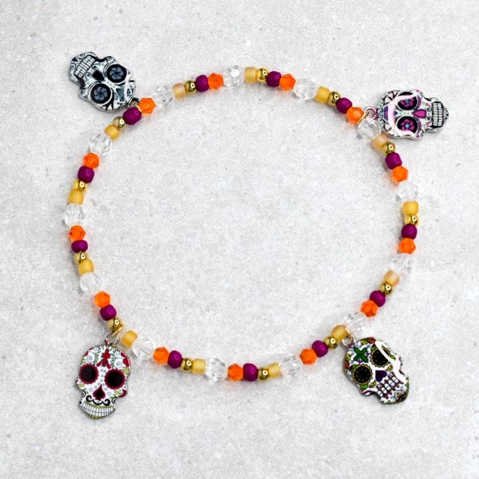A Day of the Dead themed bracelet. The bracelet features a clear beaded strand with orange, purple, and gold beads. Four silver skull charms with colorful details are evenly spaced around the bracelet.