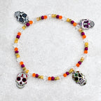 A Day of the Dead themed bracelet. The bracelet features a clear beaded strand with orange, purple, and gold beads. Four silver skull charms with colorful details are evenly spaced around the bracelet.