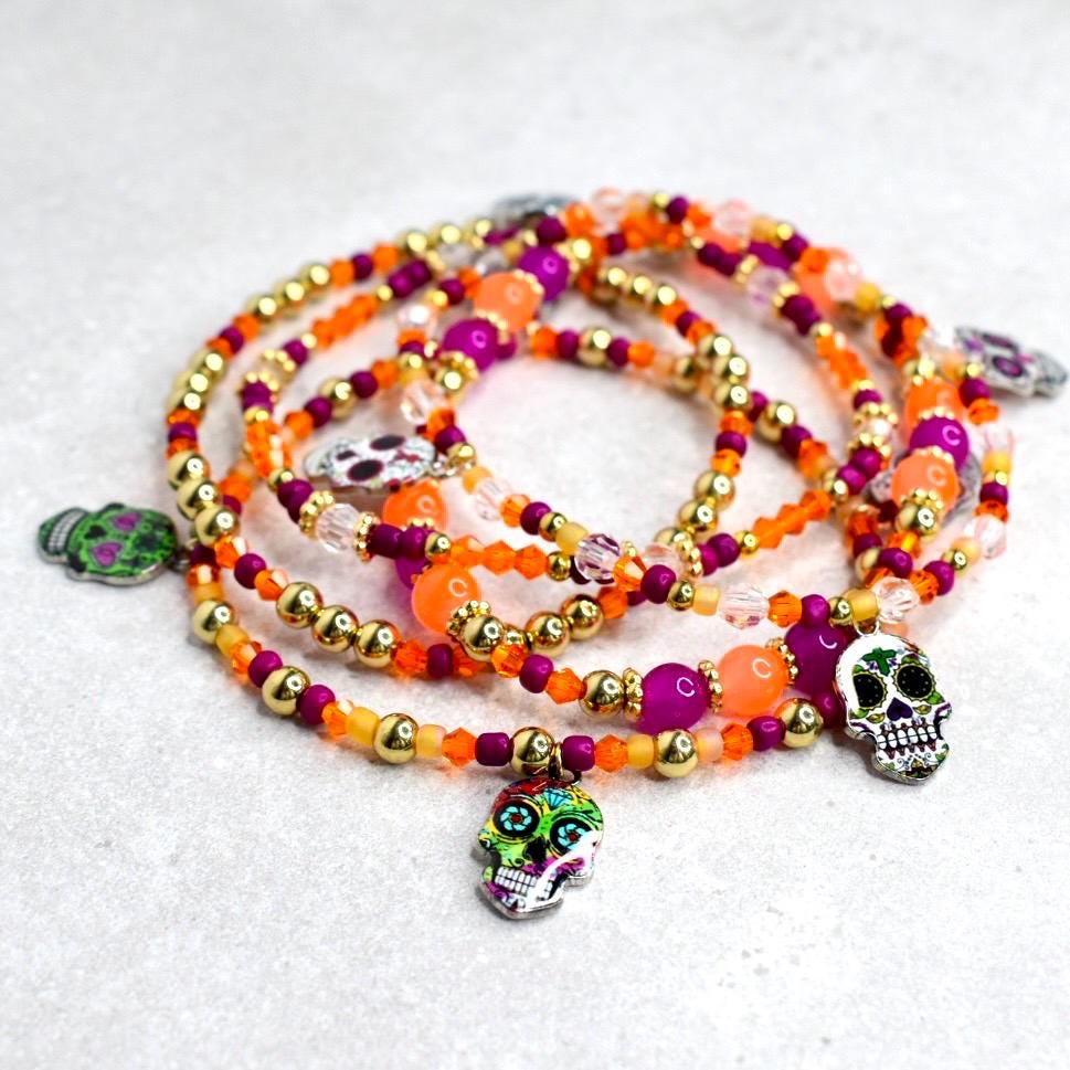 A collection of beaded bracelets in shades of orange, gold, and purple. Each bracelet features a variety of beads and is adorned with skull charms in various colors. The bracelets are arranged on a light gray surface.