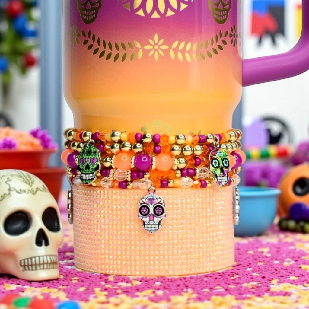 A Day of the Dead themed Stanley with a purple handle. The tumbler has a gradient design of orange, yellow, and purple. A beaded bracelet with skull charms is wrapped around the tumbler. The bracelet features gold, orange, and purple beads.