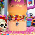 A Day of the Dead themed Stanley with a purple handle. The tumbler has a gradient design of orange, yellow, and purple. A beaded bracelet with skull charms is wrapped around the tumbler. The bracelet features gold, orange, and purple beads.