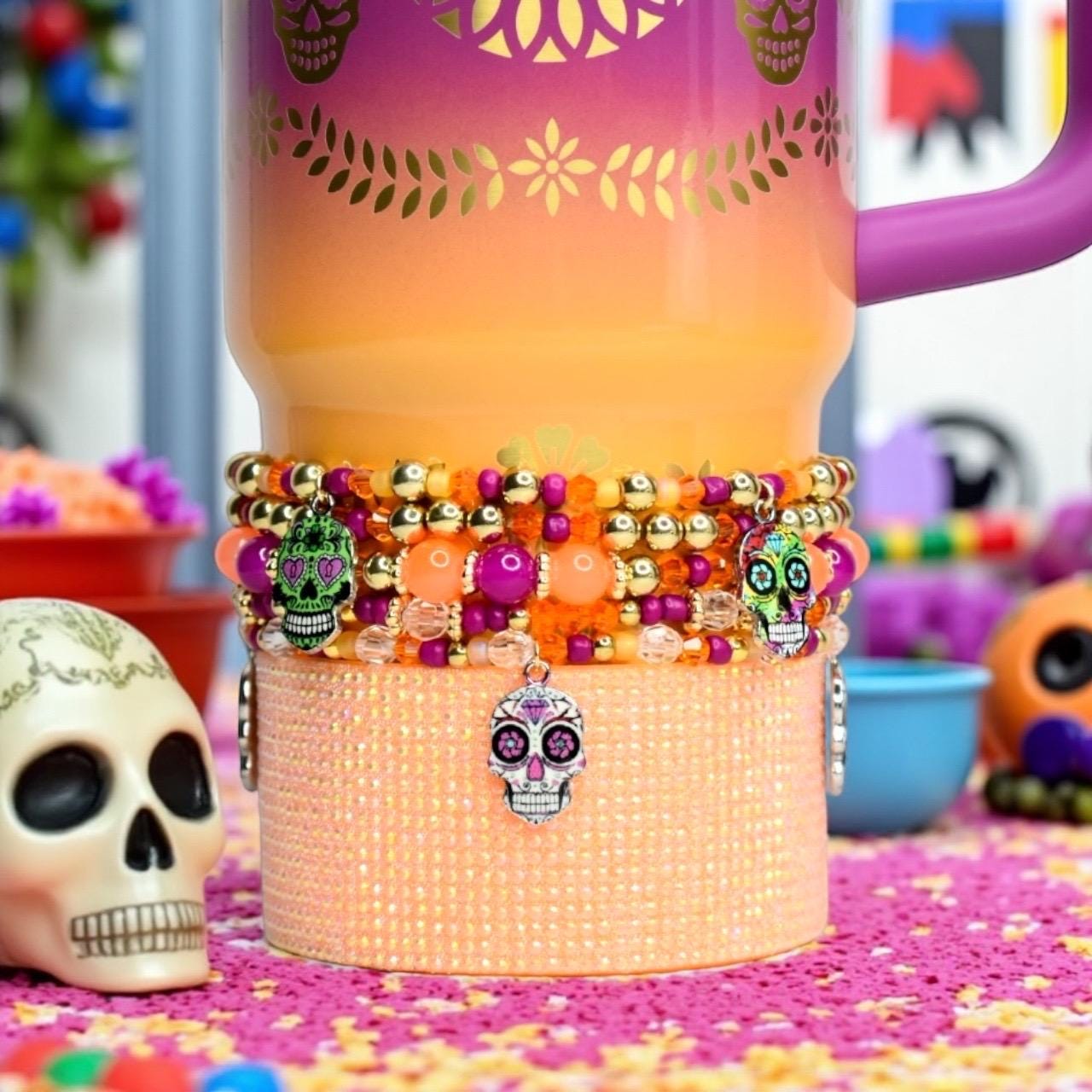 A Day of the Dead themed Stanley with a purple handle. The tumbler has a gradient design of orange, yellow, and purple. A beaded bracelet with skull charms is wrapped around the tumbler. The bracelet features gold, orange, and purple beads.