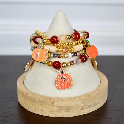 Stacked bracelets with orange pumpkin charms, gold beads, and various other beads in shades of red, cream, and gold. The bracelets are displayed on a white cone-shaped stand with a wooden base.