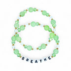 Two beaded bracelets with green and clear beads, and gold accents. The bottom bracelet spells out the word "BREATHE" in white letter beads. The bracelets are on a white background.