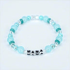 A beaded bracelet featuring light blue and clear beads. The bracelet has the word "MAMA" spelled out in black and white letter beads. The bracelet is a delicate piece of jewelry.