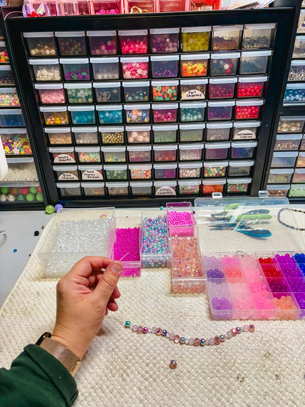 A craft station with a variety of beads in clear containers, organized in a black frame. Several open containers hold beads in pink, blue, and clear colors. A partially completed beaded bracelet is visible.