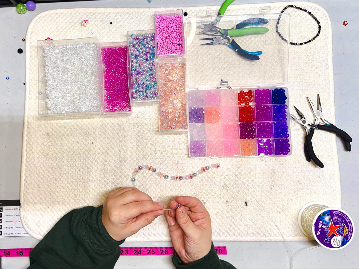 A craft setup with various beads in clear containers, a divided container of beads, and jewelry-making tools. A partially completed beaded bracelet is visible, along with a spool of string. The color palette includes pinks, purples, and clear beads.