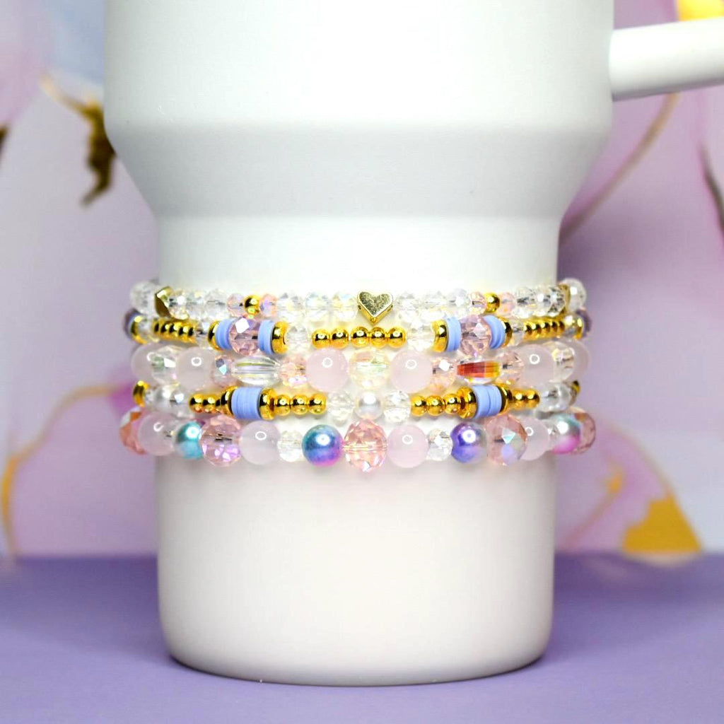 Stack of beaded bracelets in shades of pink, blue, and gold. The bracelets feature clear, pink, and iridescent beads, with gold accents and a small heart charm. The bracelets are stacked on a white mug.
