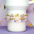 Stack of beaded bracelets in shades of pink, blue, and gold. The bracelets feature clear, pink, and iridescent beads, with gold accents and a small heart charm. The bracelets are stacked on a white mug.