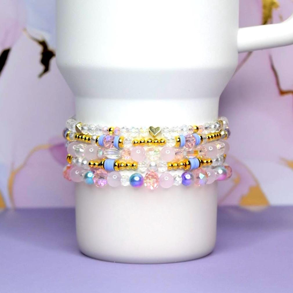 Stack of beaded bracelets in shades of pink, blue, gold, and clear. The bracelets are adorned with small beads, gold accents, and a heart charm. The bracelets are stacked on a white Stanley tumbler.
