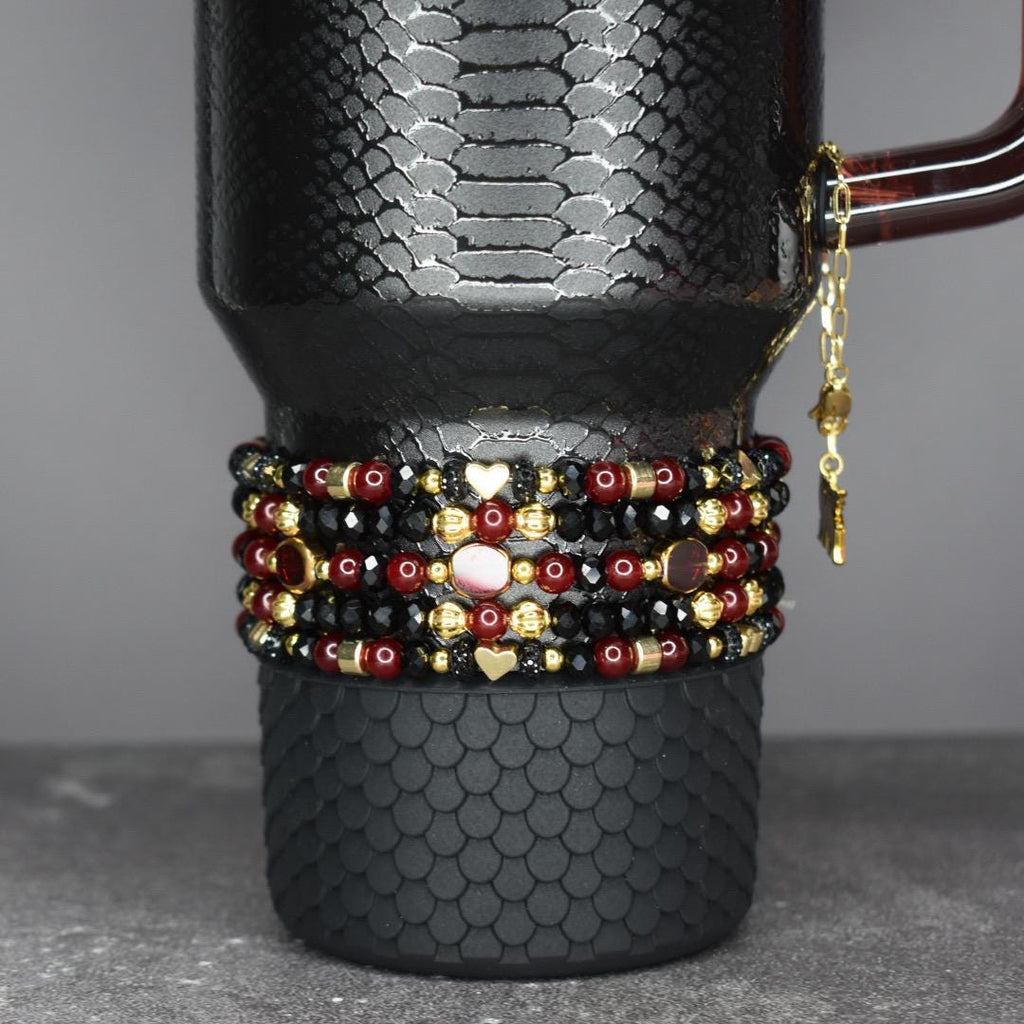 A black travel mug with a textured pattern, adorned with multiple beaded bracelets in red, black, and gold. The bracelets feature heart and star charms. A gold chain with charms hangs from the mug's handle.