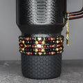 A black travel mug with a textured pattern, adorned with multiple beaded bracelets in red, black, and gold. The bracelets feature heart and star charms. A gold chain with charms hangs from the mug's handle.