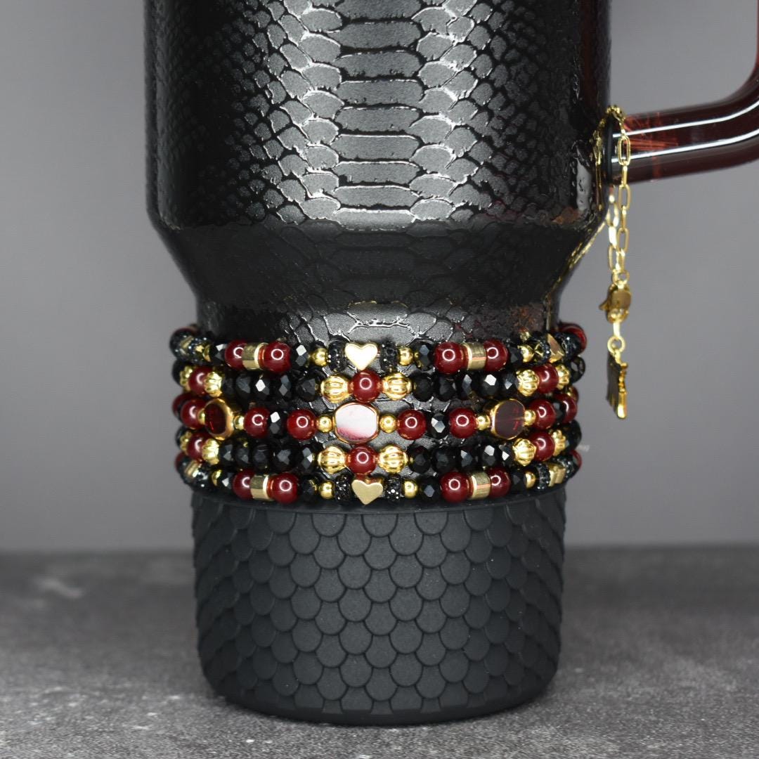 A black travel mug with a textured pattern, adorned with multiple beaded bracelets in red, black, and gold. The bracelets feature heart and star charms. A gold chain with charms hangs from the mug's handle.