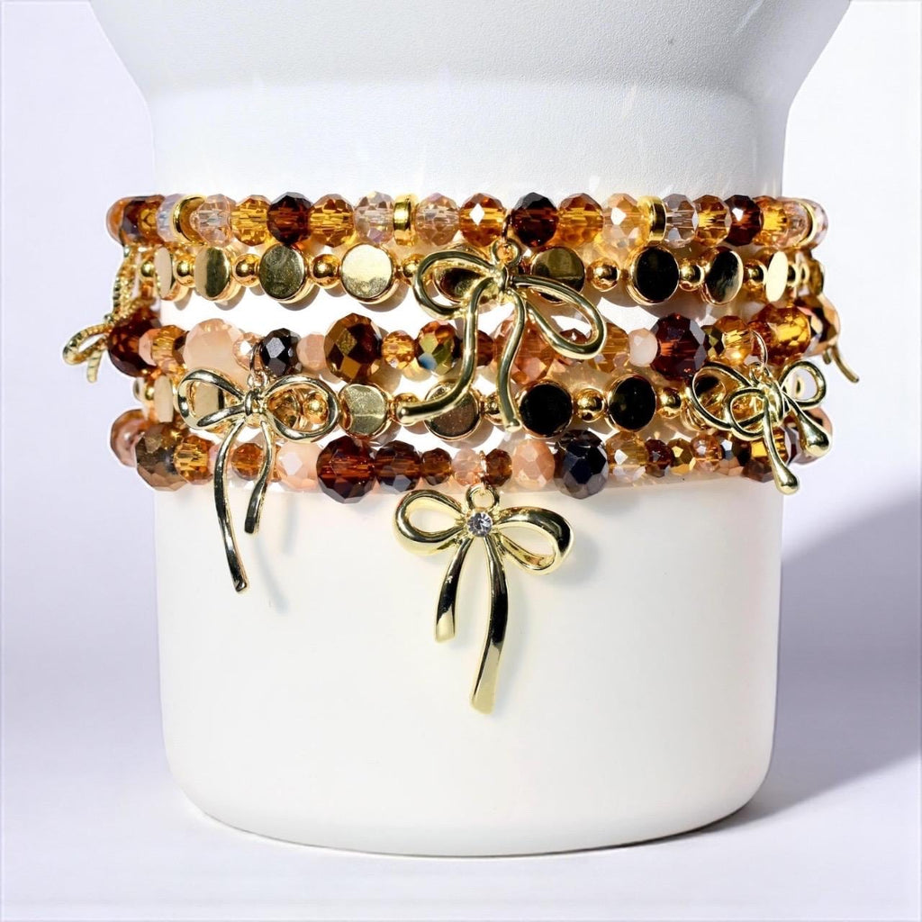 A stack of five beaded bracelets featuring faceted crystals in shades of brown, amber, and clear, interspersed with gold-toned metallic discs and small beads. Each bracelet is accented with a polished gold-toned metal bow charm.