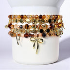 A stack of five beaded bracelets featuring faceted crystals in shades of brown, amber, and clear, interspersed with gold-toned metallic discs and small beads. Each bracelet is accented with a polished gold-toned metal bow charm.