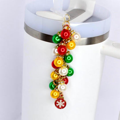 A white Stanley tumbler with a Christmas-themed charm. The charm features a cluster of red, green, yellow, and white pearl beads. A small red charm with a white snowflake hangs at the bottom. The charm is attached to the tumbler's lid with a gold chain.