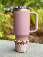 A mauve-colored tumbler with a handle and a clear lid with a gold rim. The base is decorated with a scale-like pattern. A stack of beaded bracelets in shades of pink, gold, and burgundy adorns the base.