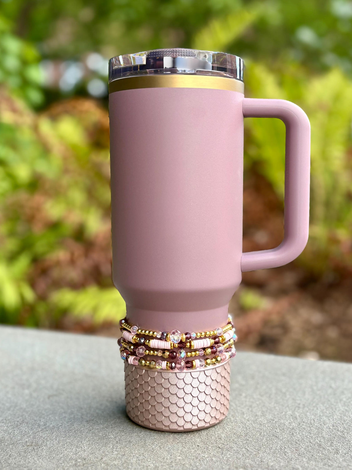 A mauve-colored tumbler with a handle and a clear lid with a gold rim. The base is decorated with a scale-like pattern. A stack of beaded bracelets in shades of pink, gold, and burgundy adorns the base.