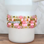 Beaded bracelet set in pink and gold tones with word beads spelling “DEFY GRAVITY,” displayed on a white tumbler.