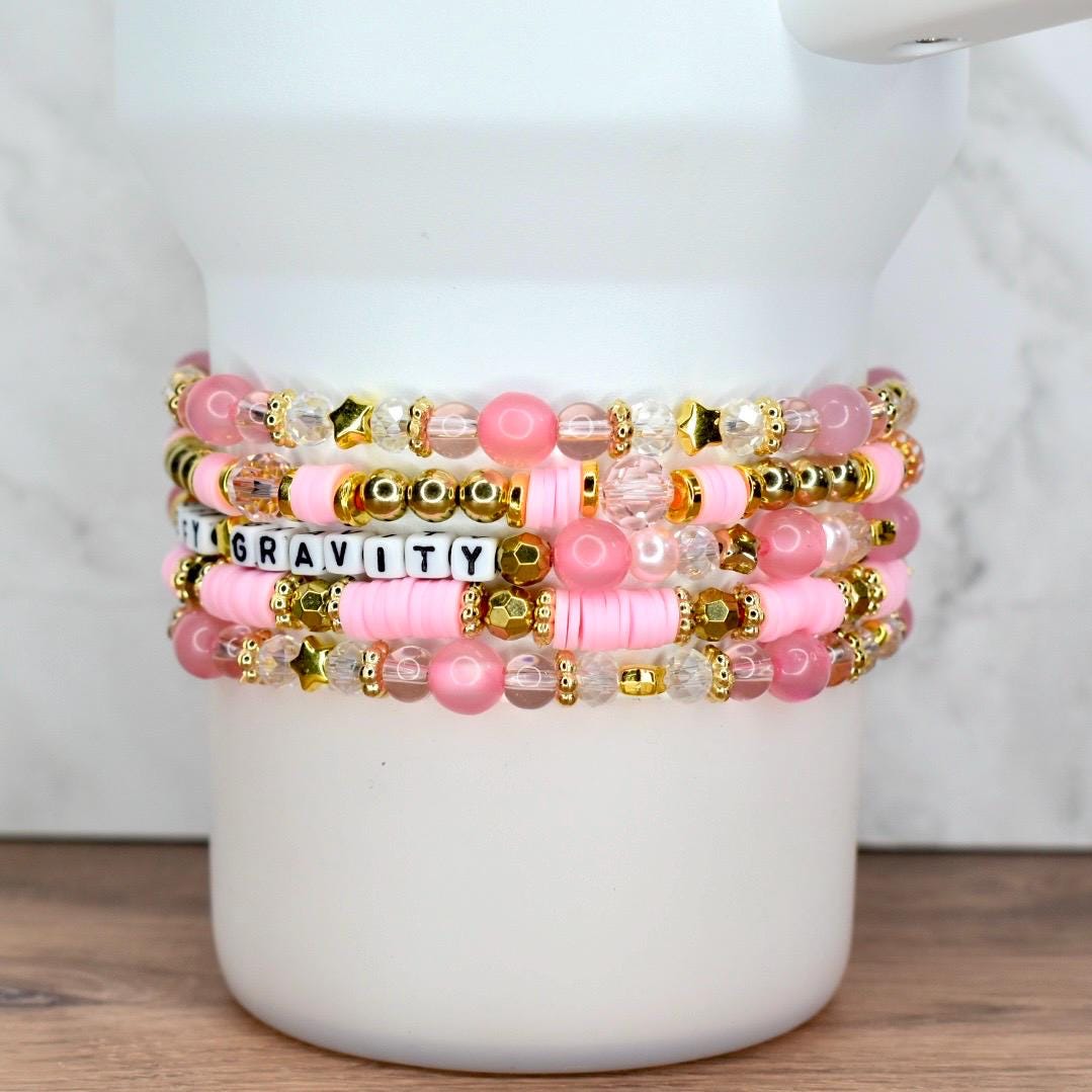 Beaded bracelet set in pink and gold tones with word beads spelling “DEFY GRAVITY,” displayed on a white tumbler.