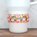 A stack of five handmade beaded bracelets in shades of pink, gold, and clear, wrapped around a white tumbler. One bracelet features letter beads spelling “DEFY GRAVITY,” while others include pearls, striped rounds, faceted beads, and gold star accents.