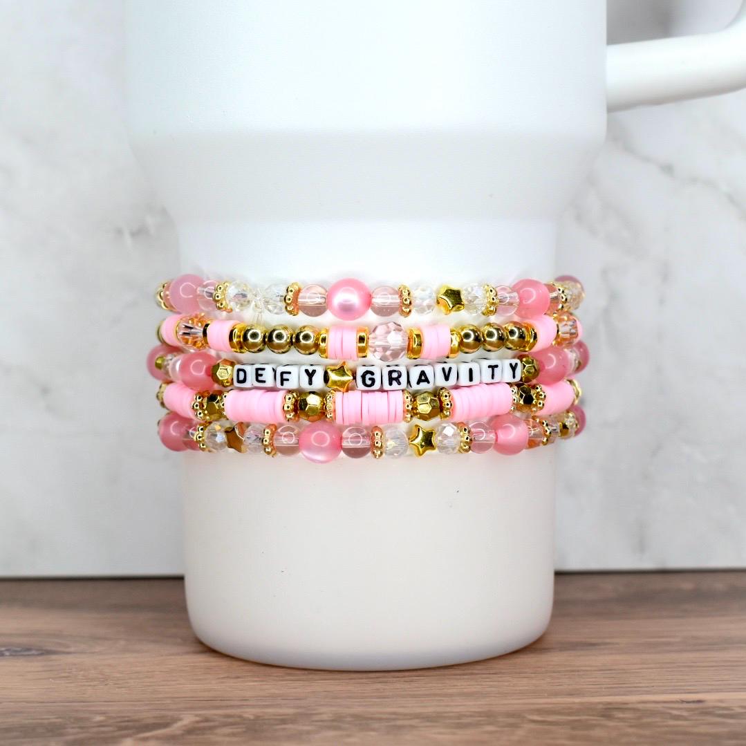 A stack of five handmade beaded bracelets in shades of pink, gold, and clear, wrapped around a white tumbler. One bracelet features letter beads spelling “DEFY GRAVITY,” while others include pearls, striped rounds, faceted beads, and gold star accents.