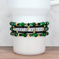 Five-bracelet stack in green, black and gold featuring letter beads spelling ‘CHANGED FOR GOOD’ with jade beads, black crystals, and gold star accents, displayed on white Stanley cup