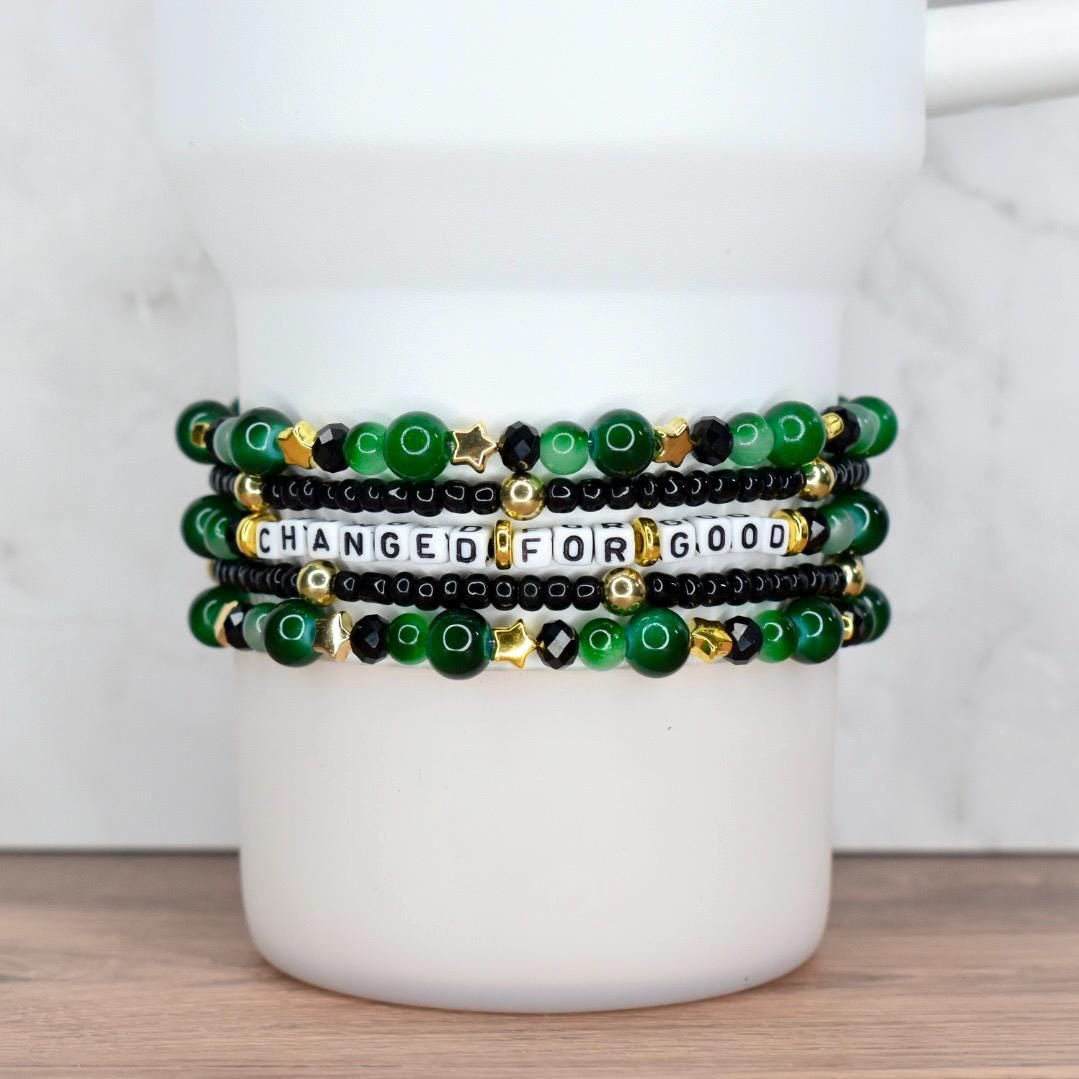 Five-bracelet stack in green, black and gold featuring letter beads spelling ‘CHANGED FOR GOOD’ with jade beads, black crystals, and gold star accents, displayed on white Stanley cup