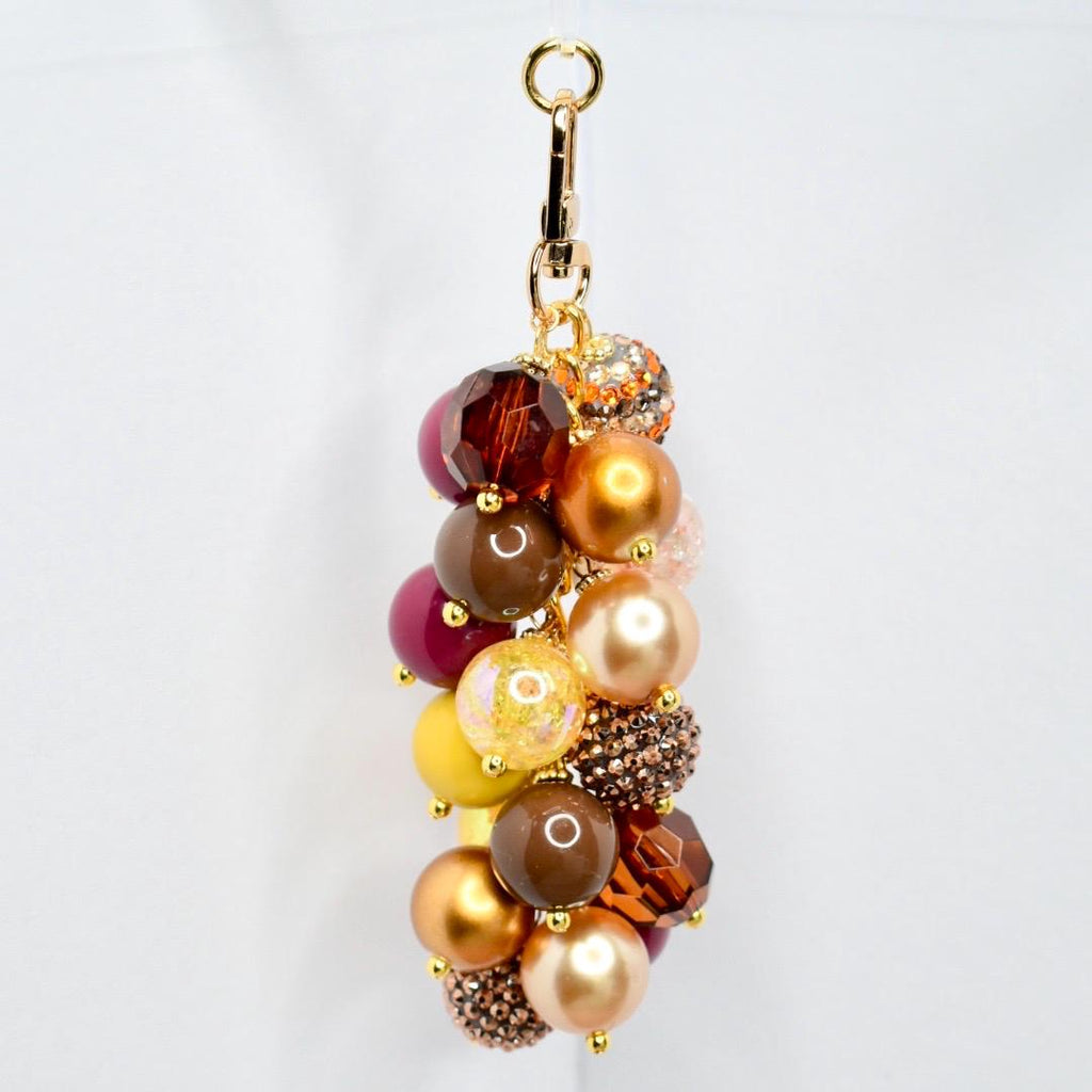 A beaded keychain with a gold-tone clasp. The keychain features a cluster of various sized beads in shades of brown, gold, yellow, and burgundy. Some beads have a sparkly, textured finish. The keychain is designed to be attached to keys or a bag.