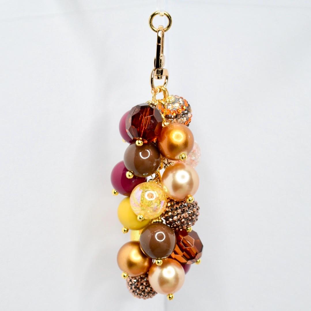 A beaded keychain with a gold-tone clasp. The keychain features a cluster of various sized beads in shades of brown, gold, yellow, and burgundy. Some beads have a sparkly, textured finish. The keychain is designed to be attached to keys or a bag.