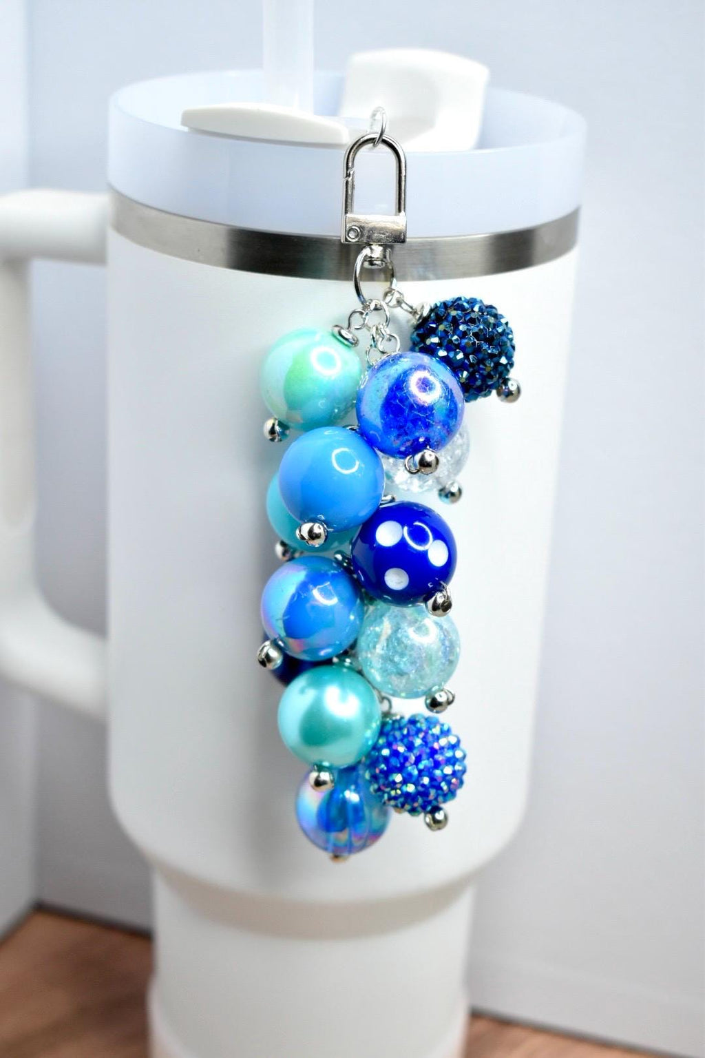 A beaded charm in shades of blue, attached to a silver clasp. The charm features various sizes of round beads, including solid, speckled, and crystal-like textures. The charm is attached to a white tumbler.