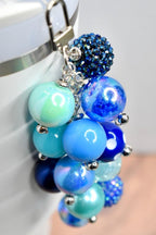 A cluster of blue and teal beads, connected by silver-tone chains and a clasp. The beads vary in size and texture, including iridescent, solid, and crystal-covered spheres. The color palette ranges from light teal to deep navy.