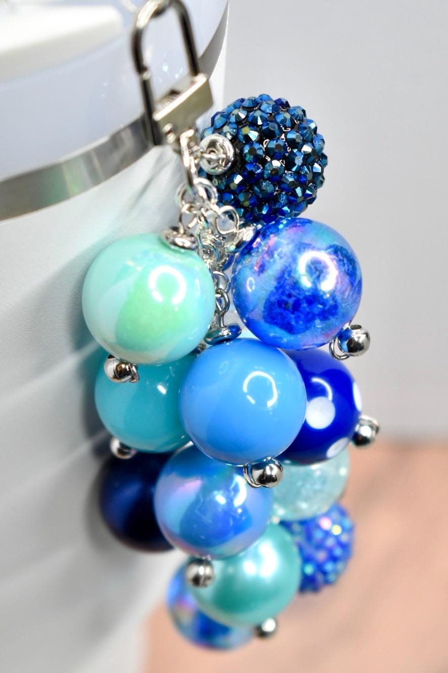 A cluster of blue and teal beads, connected by silver-tone chains and a clasp. The beads vary in size and texture, including iridescent, solid, and crystal-covered spheres. The color palette ranges from light teal to deep navy.