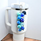 A beaded tumbler straw charm with various shades of blue and teal beads, attached to a silver clasp. The charm is hanging from a white travel mug with a silver rim and a handle. The beads vary in size and texture, creating a visually appealing accessory.