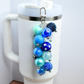 A beaded tumbler straw charm with various shades of blue beads, including solid, patterned, and crystal-like textures. The charm is attached to a silver-colored clasp and hangs next to a white tumbler.