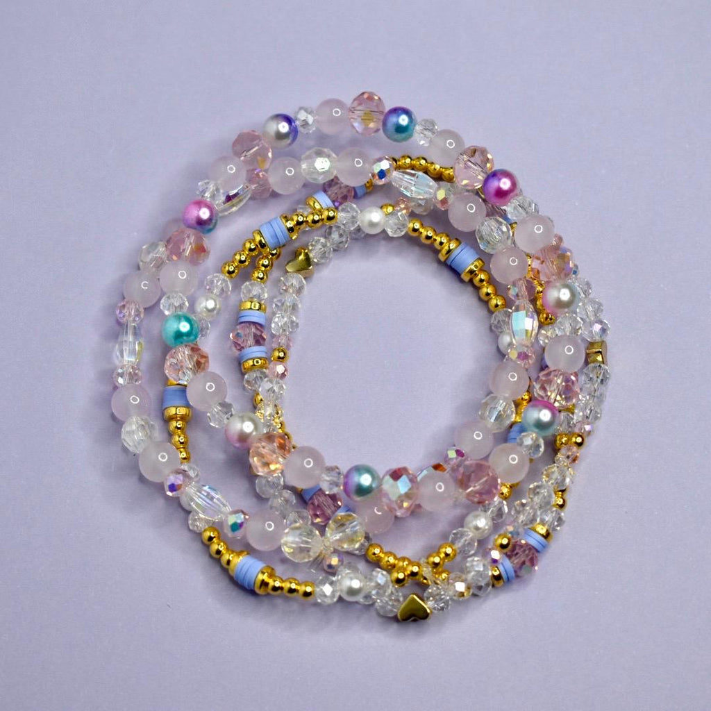 A collection of beaded bracelets in shades of pink, white, and iridescent colors. The bracelets feature a variety of beads, including round, faceted, and pearl-like beads, with gold-colored accents and light blue spacers. The bracelets are arranged in a circular pattern.