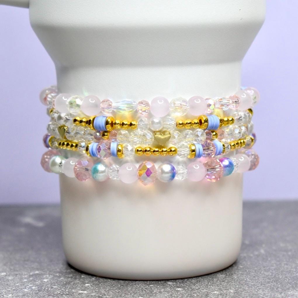 Stack of beaded bracelets in shades of pink, white, gold, and blue. The bracelets are made with various beads, including clear, iridescent, and gold-colored beads. The bracelets are stacked on a white ceramic container.