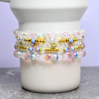 Stack of beaded bracelets in shades of pink, white, gold, and blue. The bracelets are made with various beads, including clear, iridescent, and gold-colored beads. The bracelets are stacked on a white ceramic container.