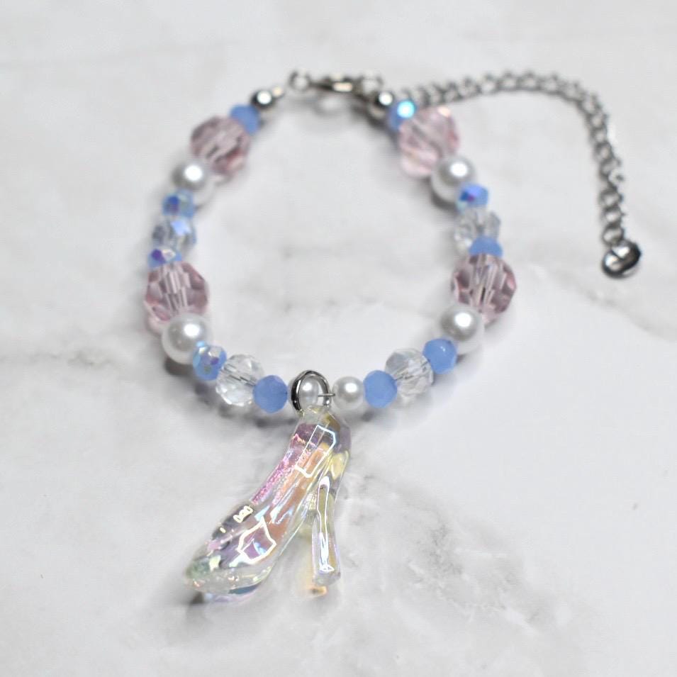 A beaded bracelet featuring pink, white, and blue beads with a clear, iridescent high-heeled shoe charm. The bracelet has a silver chain and clasp. The beads are a mix of round and faceted shapes.