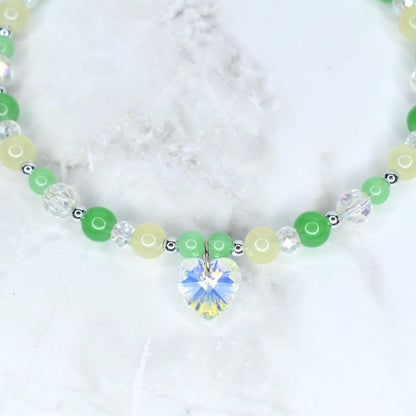 A beaded necklace featuring a heart-shaped crystal pendant. The necklace has a mix of green, yellow, and clear beads, with small silver-colored spacers. The heart pendant sparkles with multiple colors. The necklace is on a white marble surface.