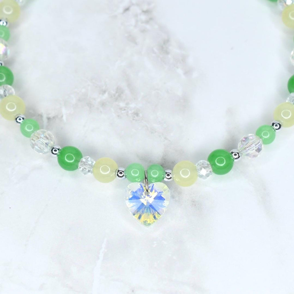 A beaded necklace featuring a heart-shaped crystal pendant. The necklace has a mix of green, yellow, and clear beads, with small silver-colored spacers. The heart pendant sparkles with multiple colors. The necklace is on a white marble surface.