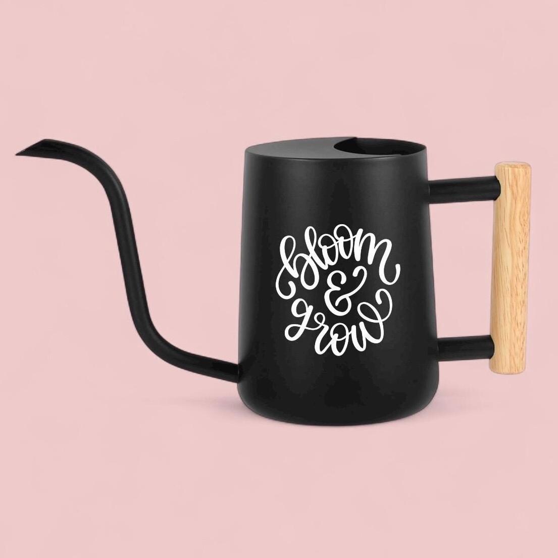 Black metal watering can with a long, curved spout and a wooden handle. The words "bloom & grow" are written in white script on the side of the can. The watering can is designed for indoor or outdoor plant care.