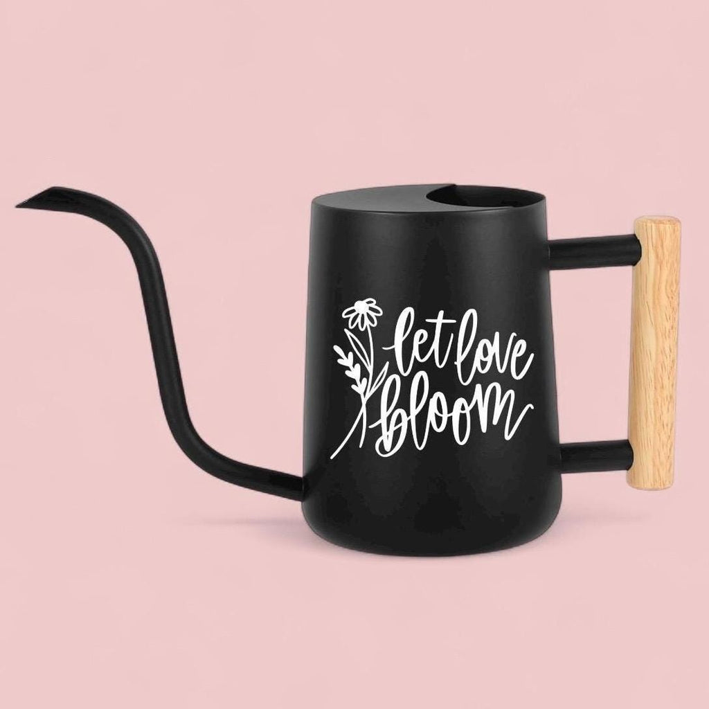A black watering can with a long, curved spout and a wooden handle. The can features white cursive text that reads 'let love bloom' with a floral design. The handle is made of light-colored wood, and the can is set against a soft, pink background
