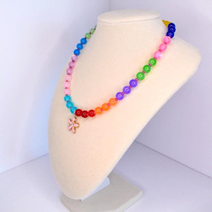 A colorful beaded necklace displayed on a mannequin. The necklace features a rainbow of beads, transitioning from blue to green, pink, orange, and red. A pink flower pendant hangs at the center. The beads are round and appear to be made of glass or a similar material.