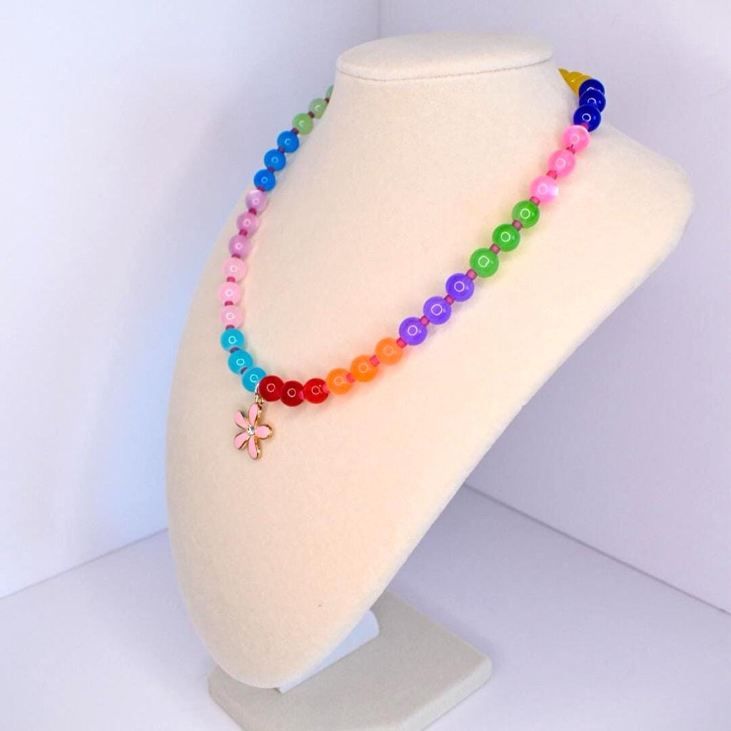 A colorful beaded necklace displayed on a mannequin. The necklace features a rainbow of beads, transitioning from blue to green, pink, orange, and red. A pink flower pendant hangs at the center. The beads are round and appear to be made of glass or a similar material.
