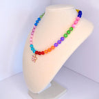 A colorful beaded necklace displayed on a mannequin. The necklace features a rainbow of beads, transitioning from blue to green, pink, orange, and red. A pink flower pendant hangs at the center. The beads are round and appear to be made of glass or a similar material.