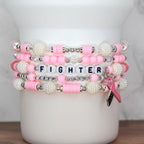 Stack of pink and white beaded bracelets. One bracelet spells "FIGHTER" with white letter beads. A pink ribbon charm hangs from one bracelet. The bracelets are displayed on a white cylindrical object.