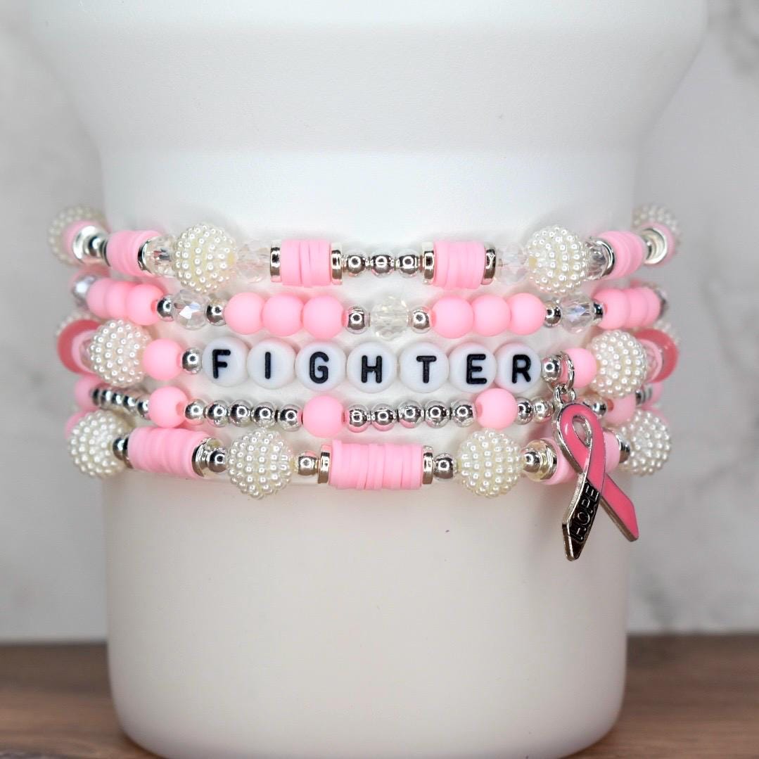 Stack of pink and white beaded bracelets. One bracelet spells "FIGHTER" with white letter beads. A pink ribbon charm hangs from one bracelet. The bracelets are displayed on a white cylindrical object.