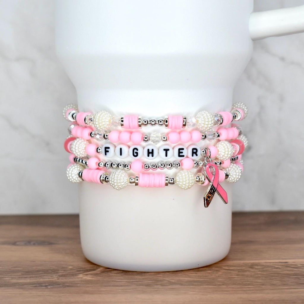 Stack of pink and white beaded bracelets with the word "FIGHTER" spelled out in letter beads. A pink ribbon charm hangs from one bracelet. The bracelets are displayed on a white mug.