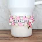 Stack of pink and white beaded bracelets with the word "FIGHTER" spelled out in letter beads. A pink ribbon charm hangs from one bracelet. The bracelets are displayed on a white mug.